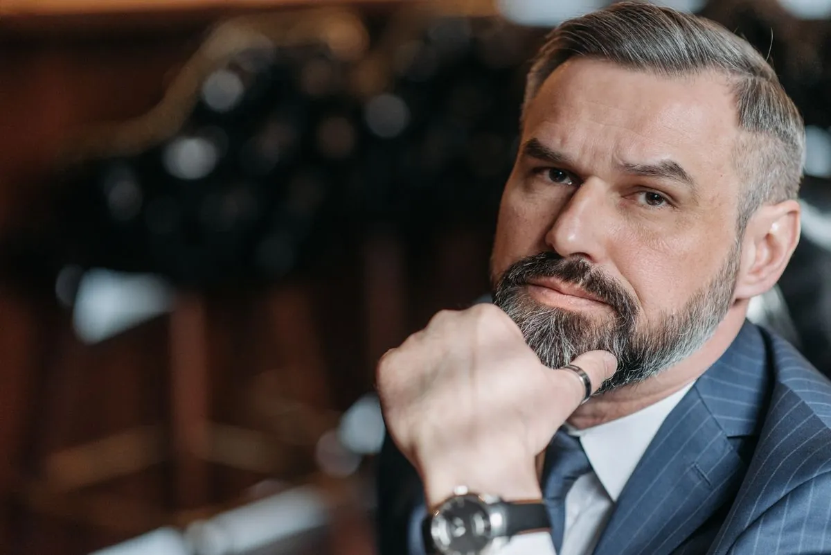 An older man with a grey beard in a dark suit, hand at his chin